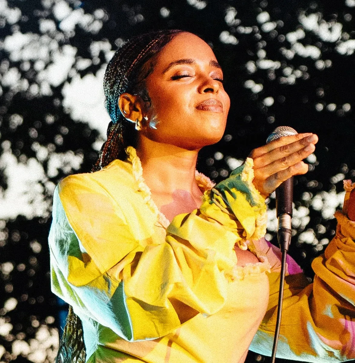 Vocalist and composer Roxanne Tataei mid-song. Her eyes are closed and face is tilted up, she is holding a microphone and wearing a yellow dress