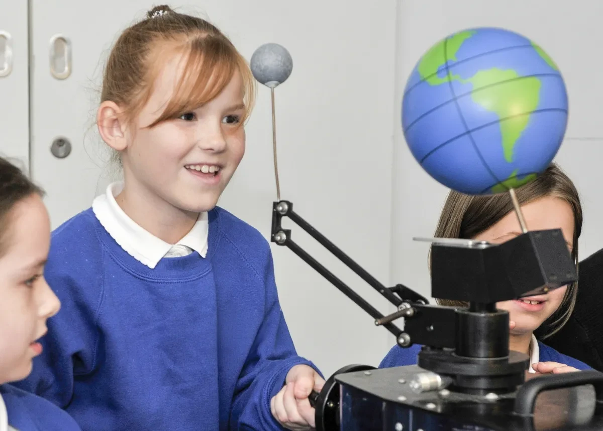 Children in school uniform interact with a model of the Earth and Moon.