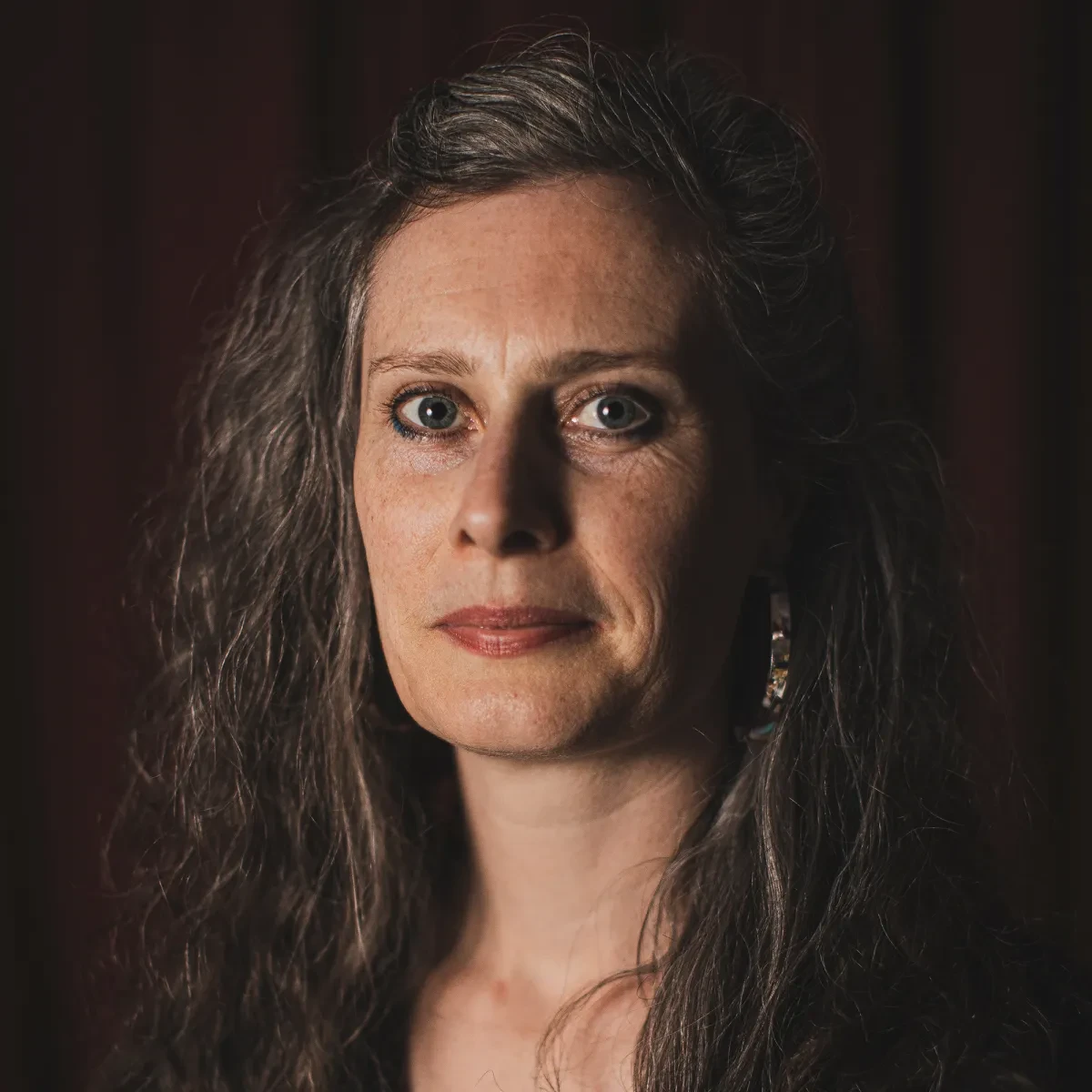 Portrait photograph of artist Brona Martin. She has long greying hair, and is lit from the side as she gazes steadily at the camera