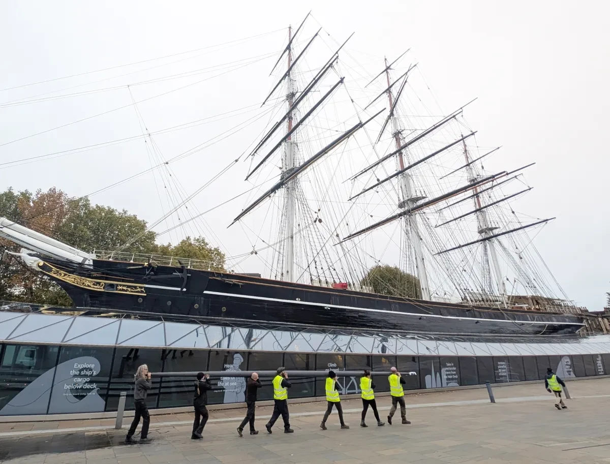 A group of people wearing high vis jackets carry a beam on their shoulders in front of the Cutty Sark ship