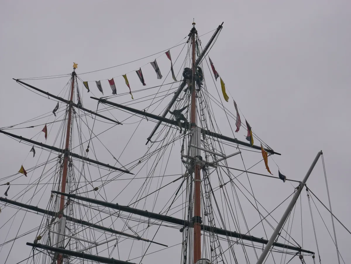 A group of shipwrights install a horizontal beam at the top of a ship's mast