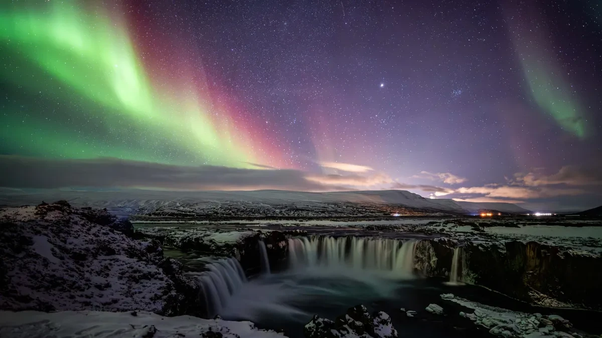 Landscape photo showing large, deep waterfall in a snowy landscape with mountains in the distance. The aurora is in the starry sky in vivid greens and purples above