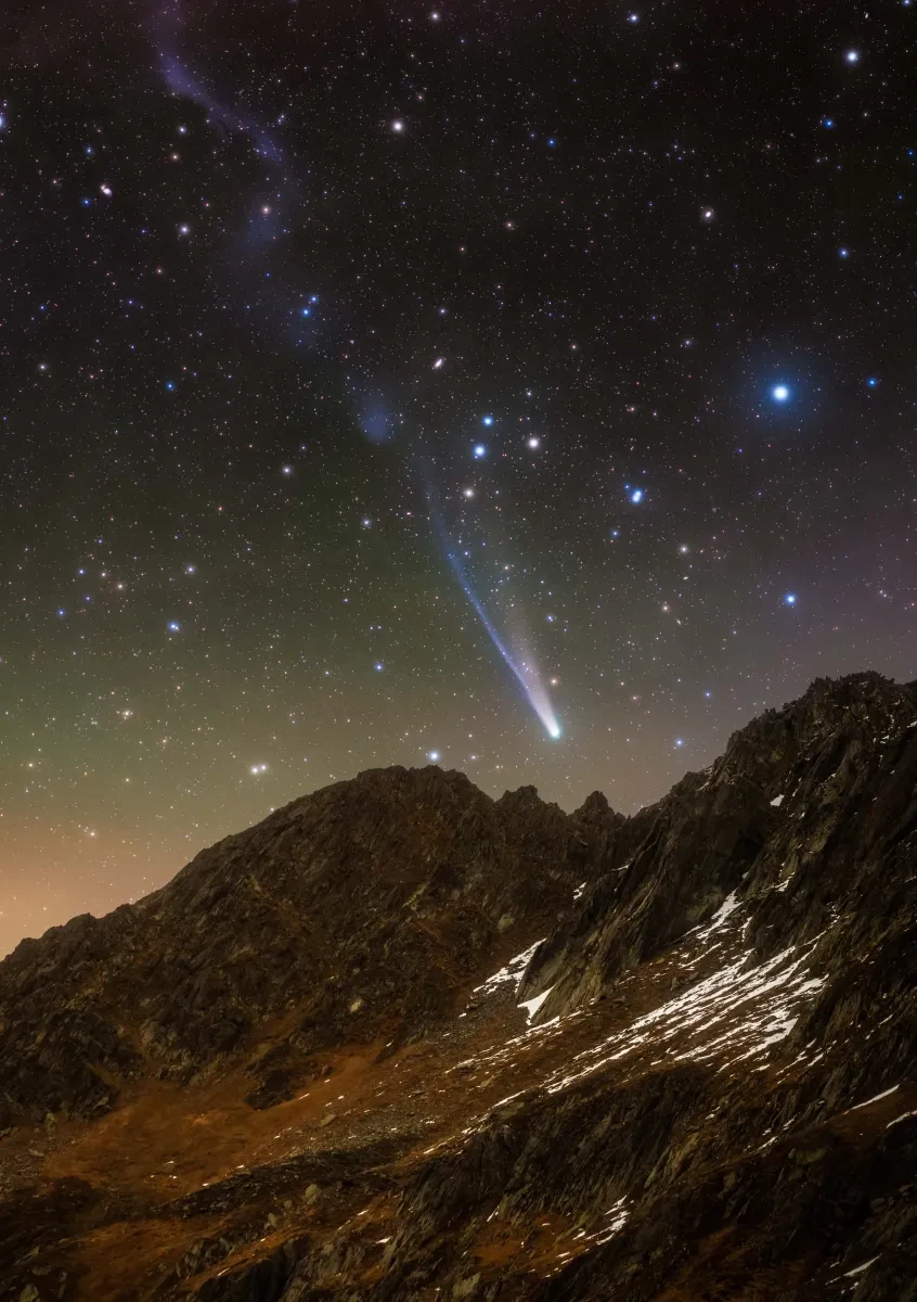 Photo of sloping mountain with a bright comet in the sky above with a white head and long blue tail. The sky is full of bright glowing stars in blues and oranges