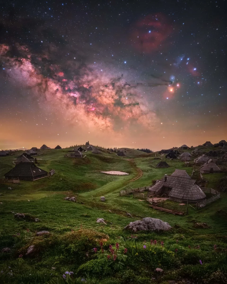 Lush green hilly landscape dotted with historic small buildings. In the sky the Milky Way runs diagonal and appears in bright purples, oranges and pinks