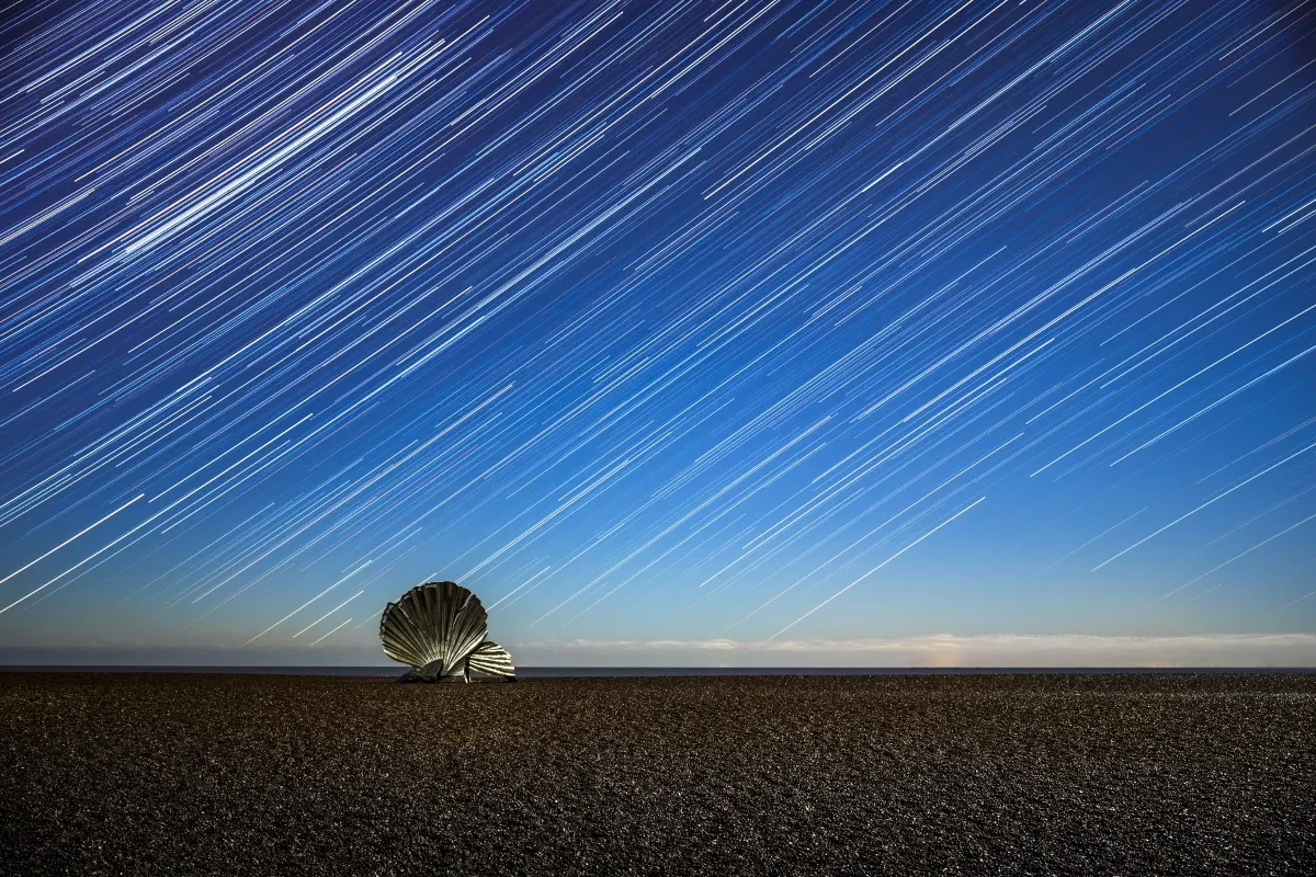 Shell sculpture on pebbly beach with star trails in the sky above and yellow glow on the horizon