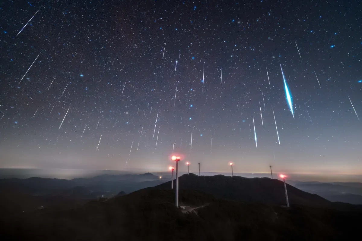 Tens of meteors falling directly downwards in the sky, one of them especially bright, over a high-up mountainous area with phone masts lit up 