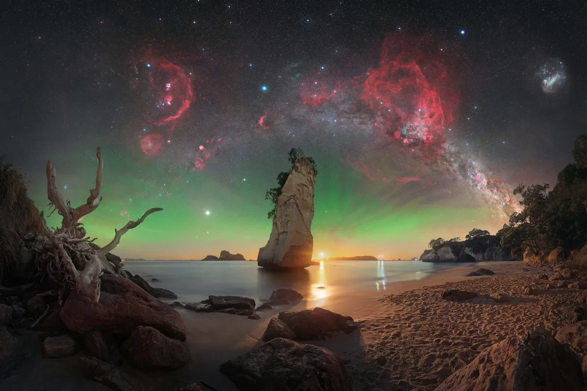 Rock formation in sea slightly off sandy beach, with green auroral glow in sky above and Milky Way arch. Vivid red nebulae stand out in the arch