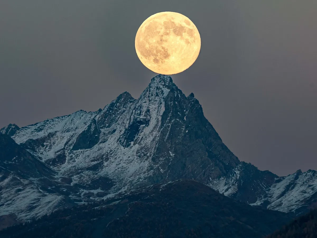 Photo of snowy mountain in low lighting, with a vivid, bright large Moon in soft yellow tone above