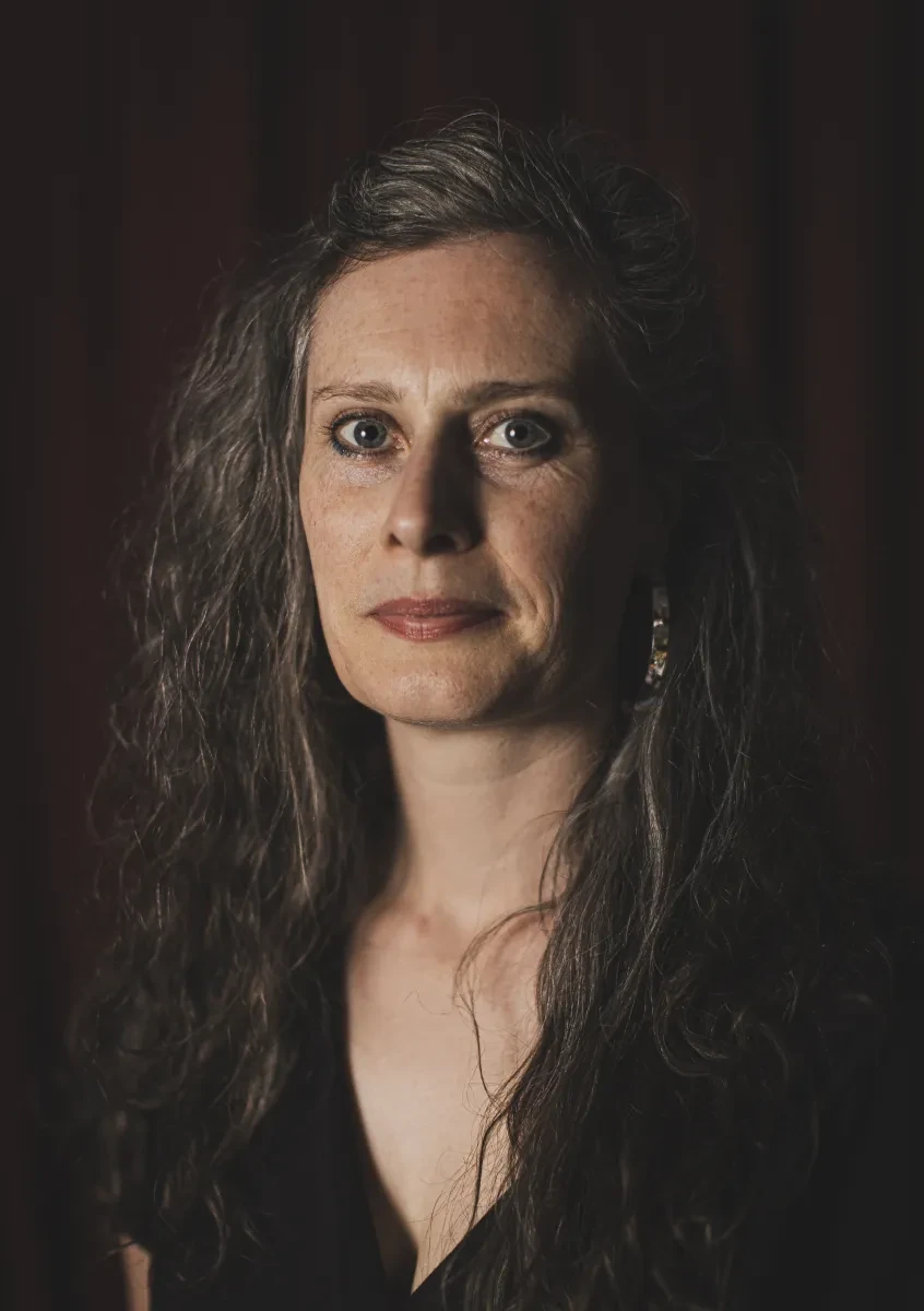 A woman with long dark hair and pale skin, wearing a dark shirt and a half-smile looks intensely at the camera. She's standing in shadow