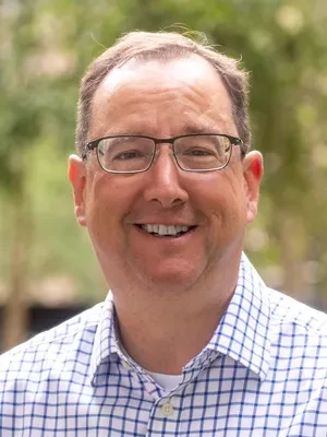 A smiling man with short brown hair, wearing a blue checked shirt and glasses