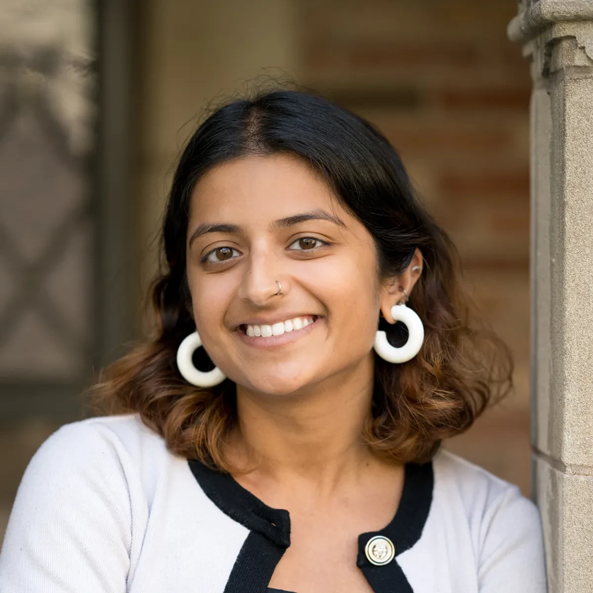 A woman with mid-length dark brown hair, wearing large white hoop earrings, smiles while leaning on a stone pillar