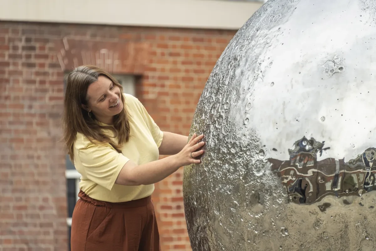 A smiling woman in a yellow t-shirt runs her hands over the craters of Luke Jerram's silvery Mirror Moon sculpture at the Royal Observatory in Greenwich