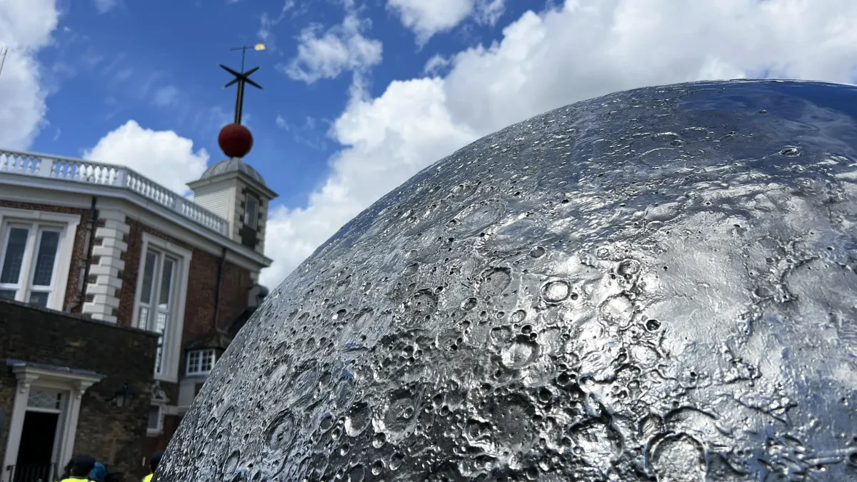 A silver Moon sculpture sits in front of an historic building with a red time ball on the roof
