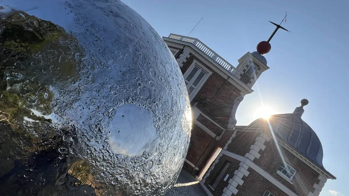 A silver sculpture of the Moon sits in front of a historic building with a red time ball on the roof. The sun shines over the sculpture