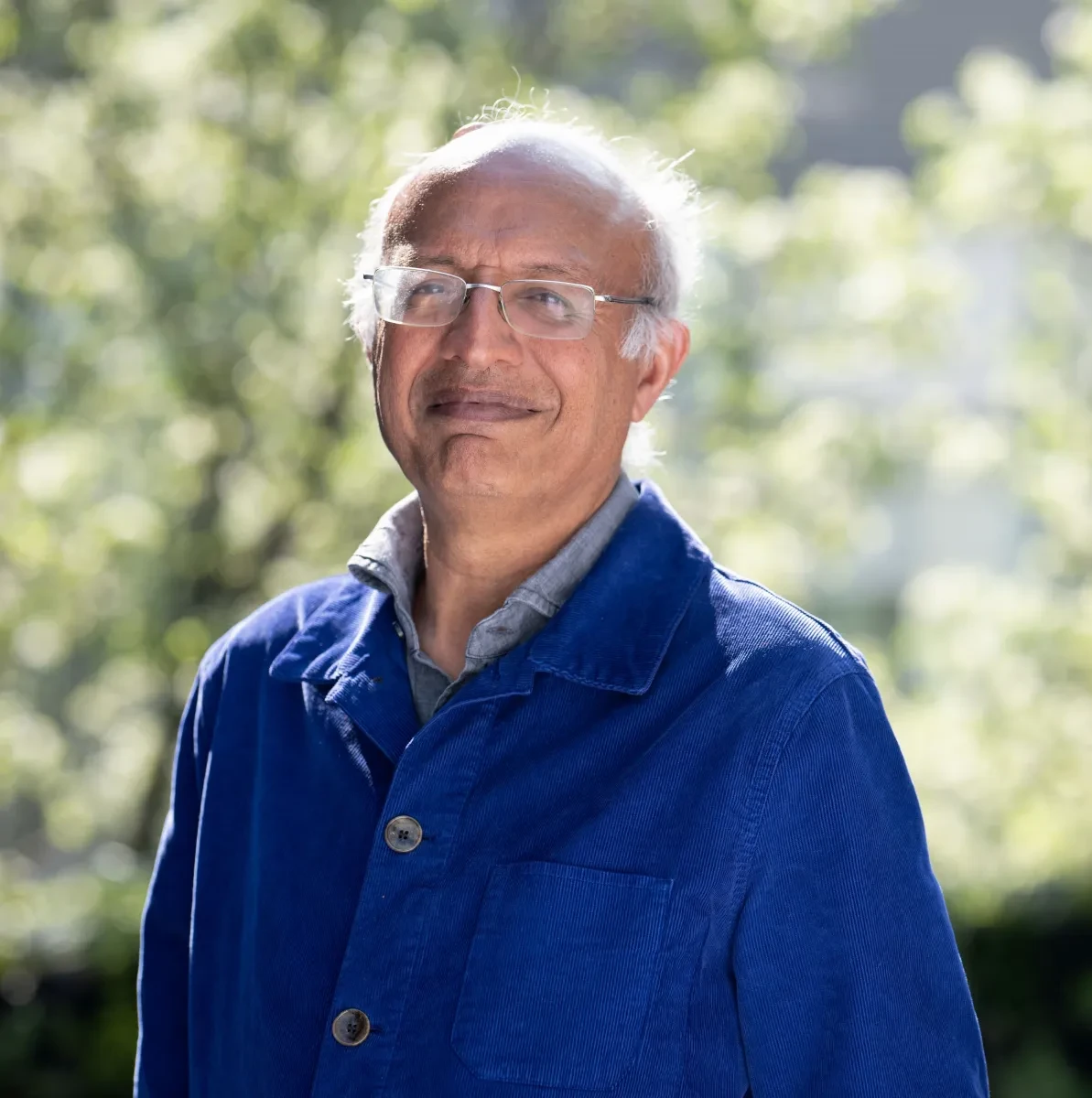 A smiling man wearing glasses and a blue jacket stands in a sunny garden