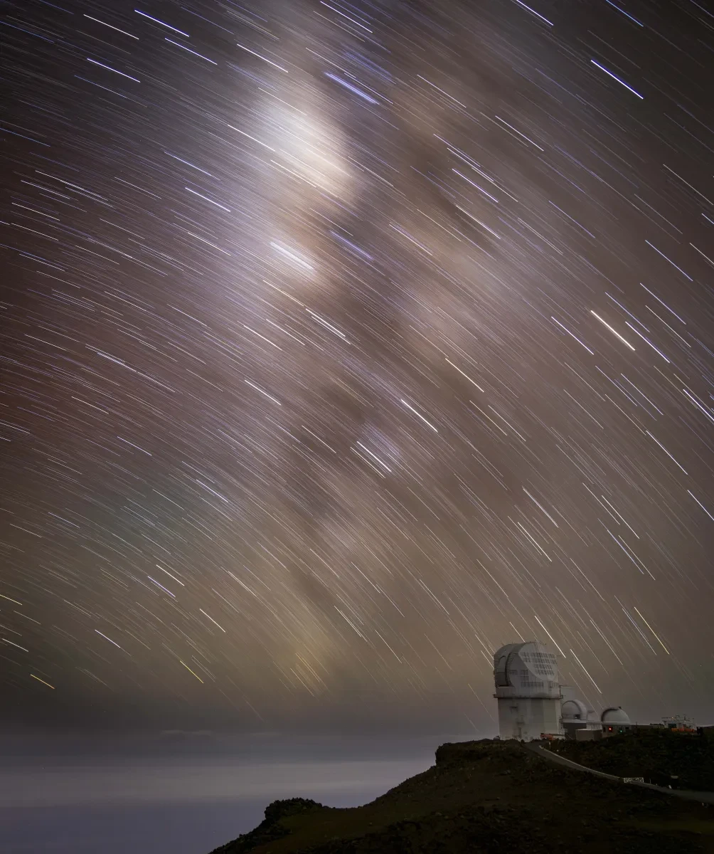 A curve of white streaks across a light-brown sky with a clifftop and a building in the foreground