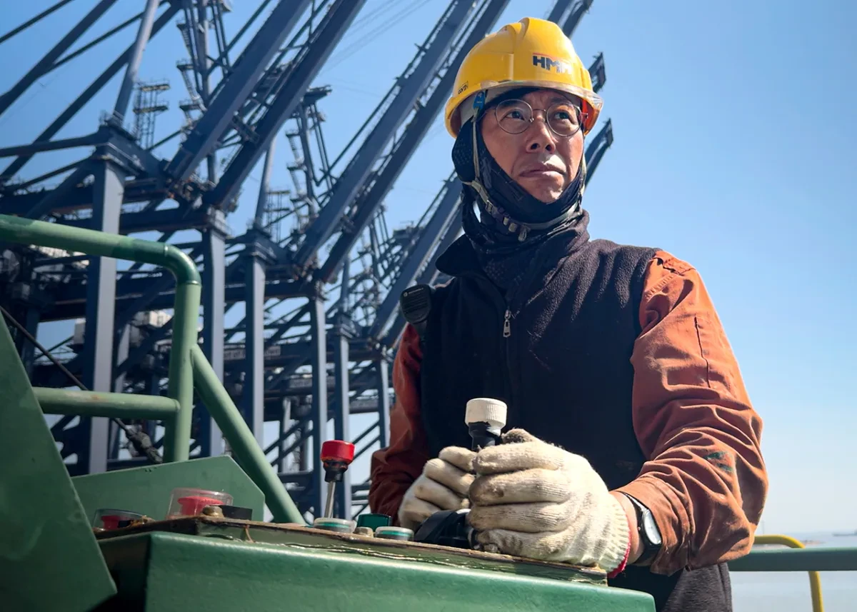 A man in orange overalls, black fleece, yellow helmet and thick gloves operators a joystick, with a view of large harbour cranes in the background
