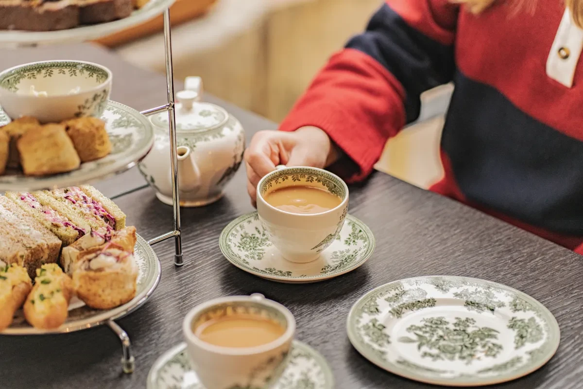 Two full teacups, a teapot, and a tiered cake stand and an empty plate on a table, with a person in a red and blue striped jersey taking hold of one of the teacups to have a drink