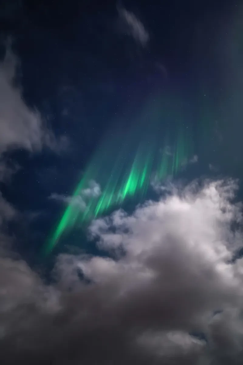 A stunning photo of vertical streaks of bright green aurorae between expanses of blue sky and fluffy white clouds