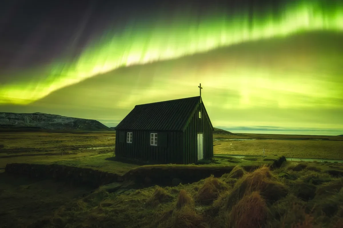 A small hut or chapel with a cross on the roof sits in a grassy landscape; the sky above is a striking mass of yellow and green aurorae framing the whole vista