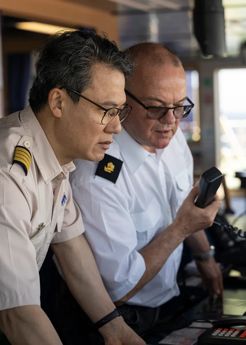 A ship's captain and a deep sea pilot discussing something on the bridge of a ship. The pilot has a phone receiver in his hand