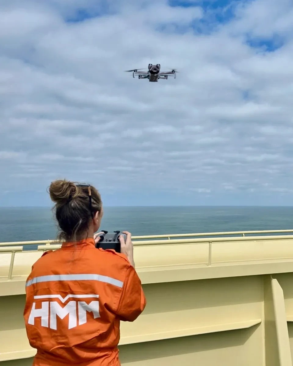 A woman operates a drone from the deck of a ship. She is wearing orange overalls with the letters 'HMM' on the back, is holding a controller and watching the drone hovering above her