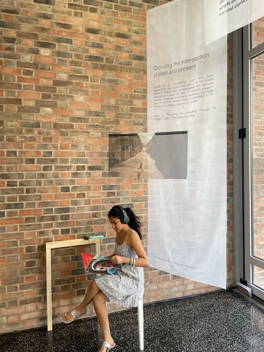 photo of a young woman sitting on a chair, with headphones on. Against a backdrop of brown/red brick wall and cotton hanging behind her.n