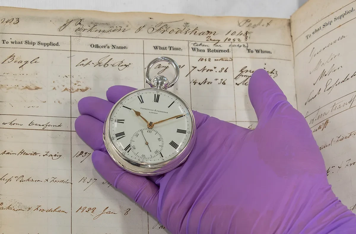 A silver and white pocket chronometer with black roman numerals on a purple gloved hand, next to an old ledger detailing the voyage of HMS Beagle