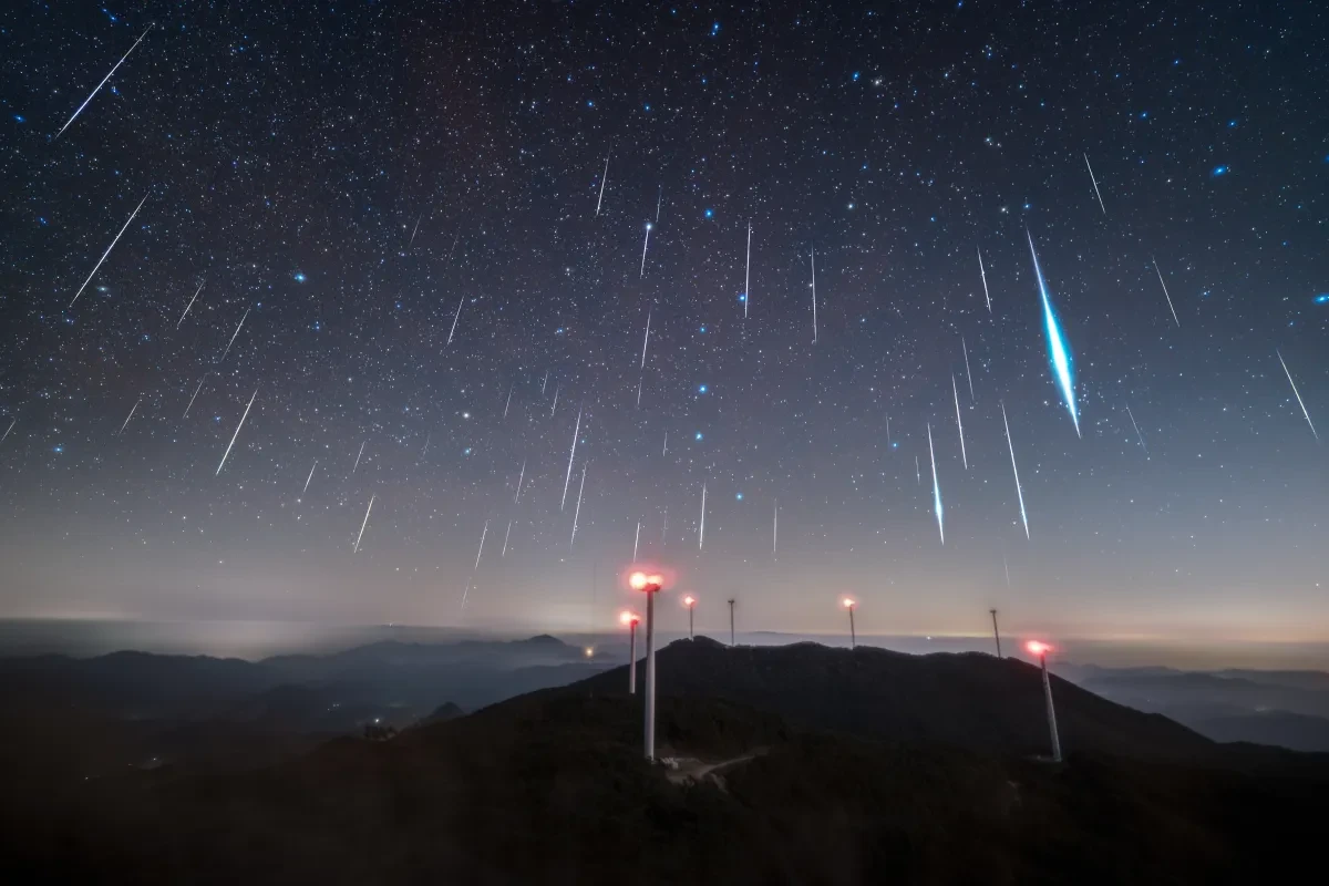 Tens of meteors falling directly downwards in the sky, one of them especially bright, over a high-up mountainous area with phone masts lit up