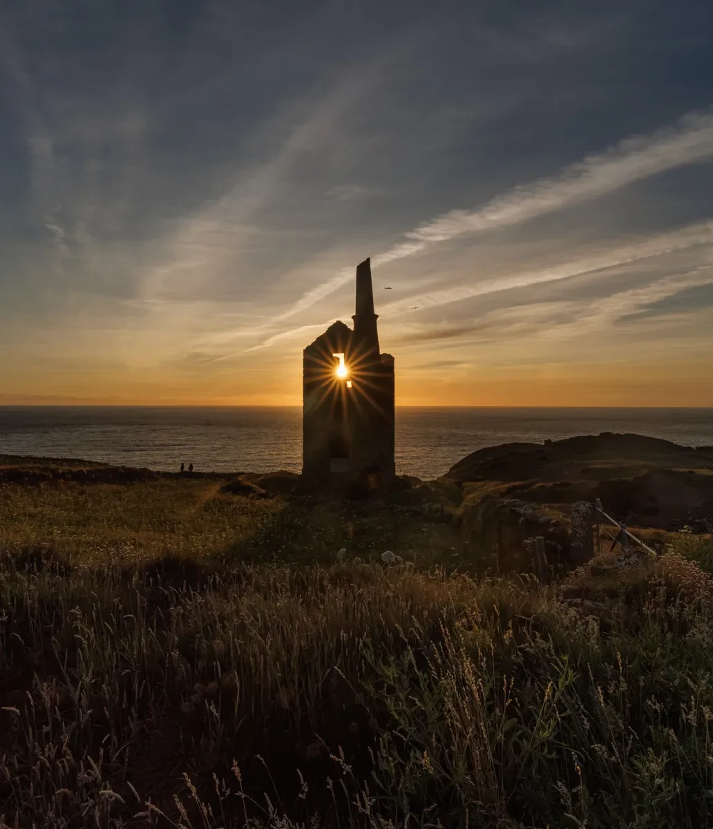 Photo of Cornish tin mine on cliff edge looking towards the sea, with the setting Sun glowing in the upper window of the ruined structure