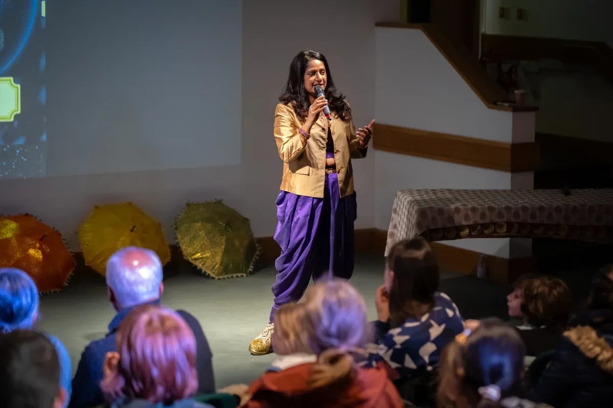 A speaker in purple trousers and a light brown jacket with long black hair talks into a microphone on stage in the National Maritime Museum's lecture theatre