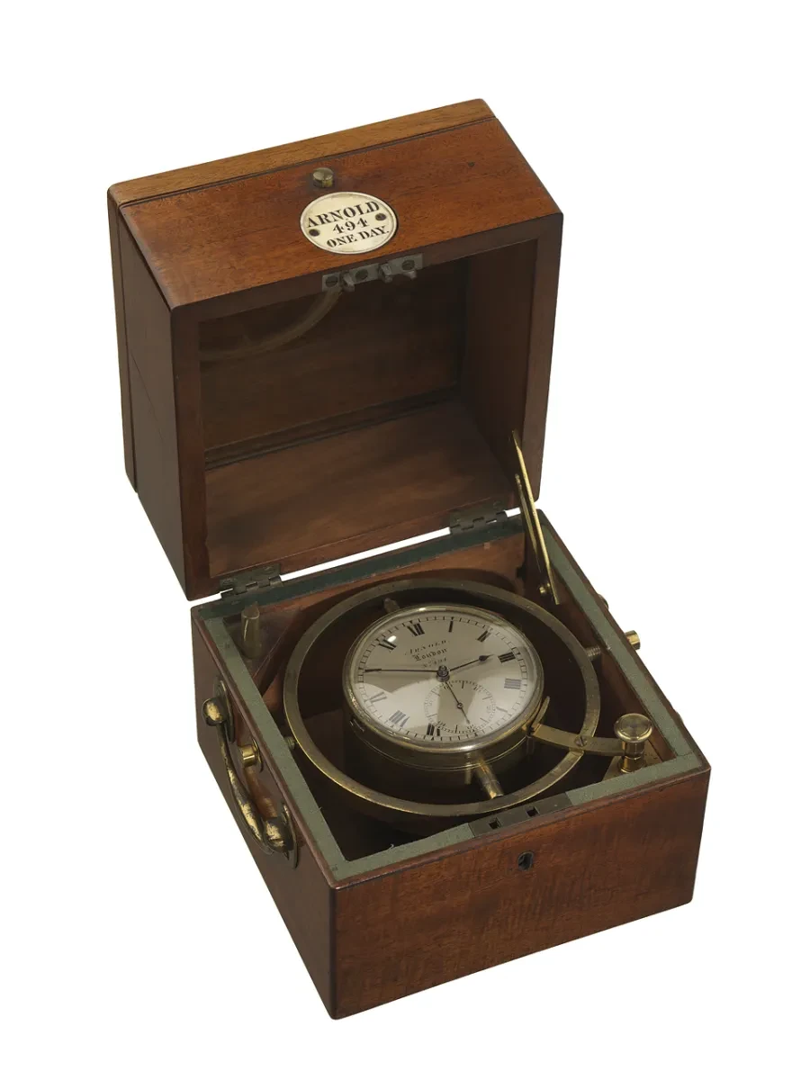 A chronometer with a silvery face surrounded by a metal bar to keep it stable, sits in a dark brown box
