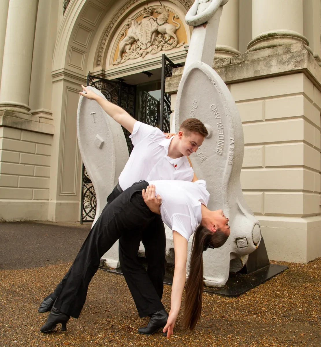 colour photo of 2 dancers, a young man holding holding a young woman as she bends backwards in front of a large white anchor in front of a cream coloured building.