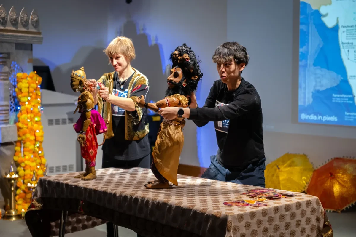 Two puppeteers stand behind a table covered in a patterned table cloth while manipulating puppets in front of a large screen in a lecture theatre at the National Maritime Museum