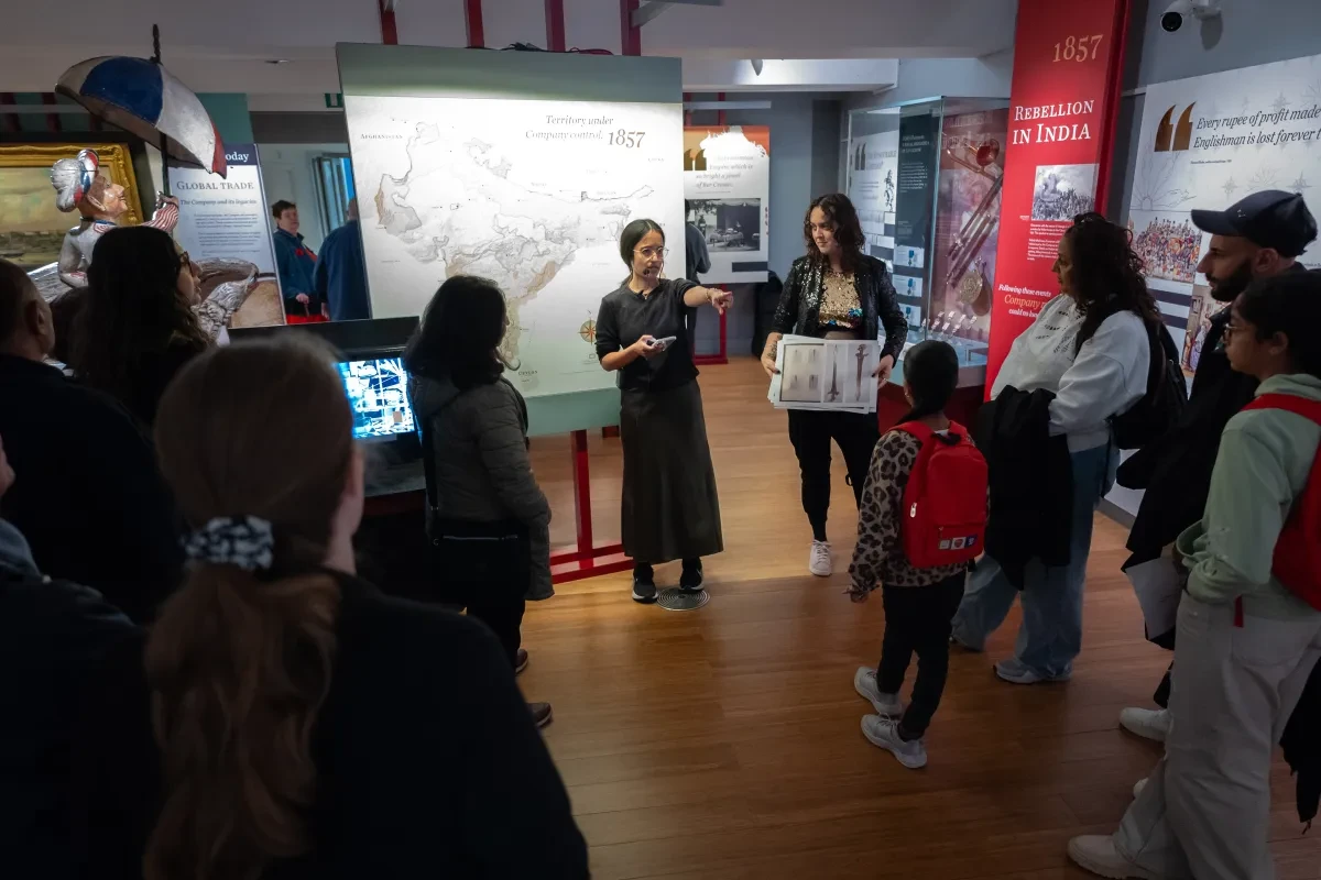 A woman gives a talk in a gallery within the National Maritime Museum while members of the public stand and listen