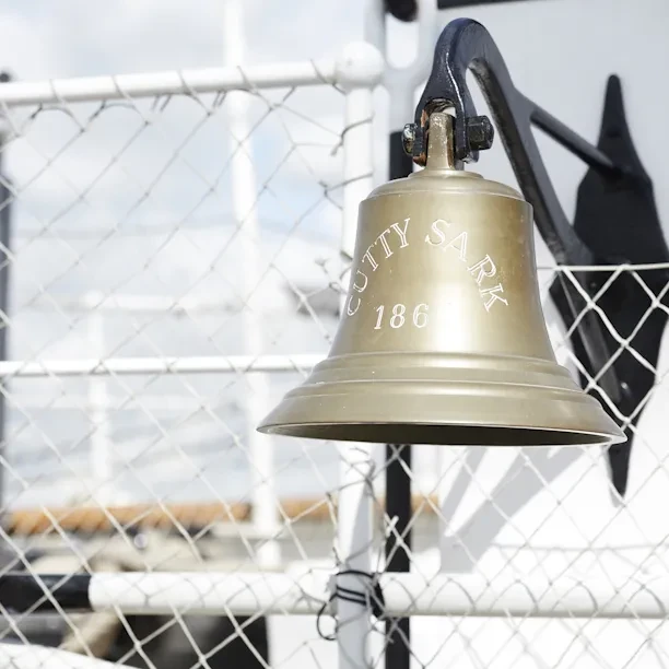 A ship's bell engraved with the name Cutty Sark is mounted on white wire fencing.
