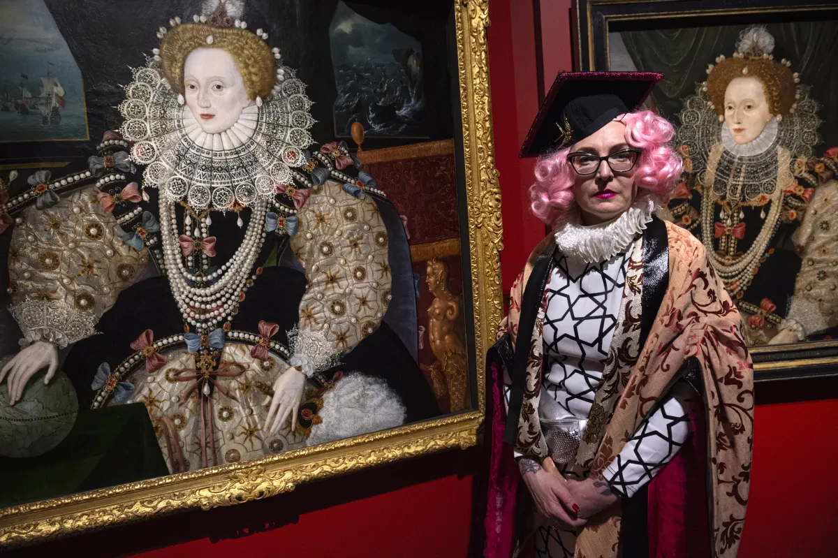 A person wearing patterned clothing, pink wig and graduation-style cap stands next to a painting of Elizabeth the first.