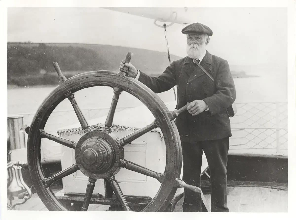 A black and white photograph of a bearded man in a flat cap steering with a large ships wheel.