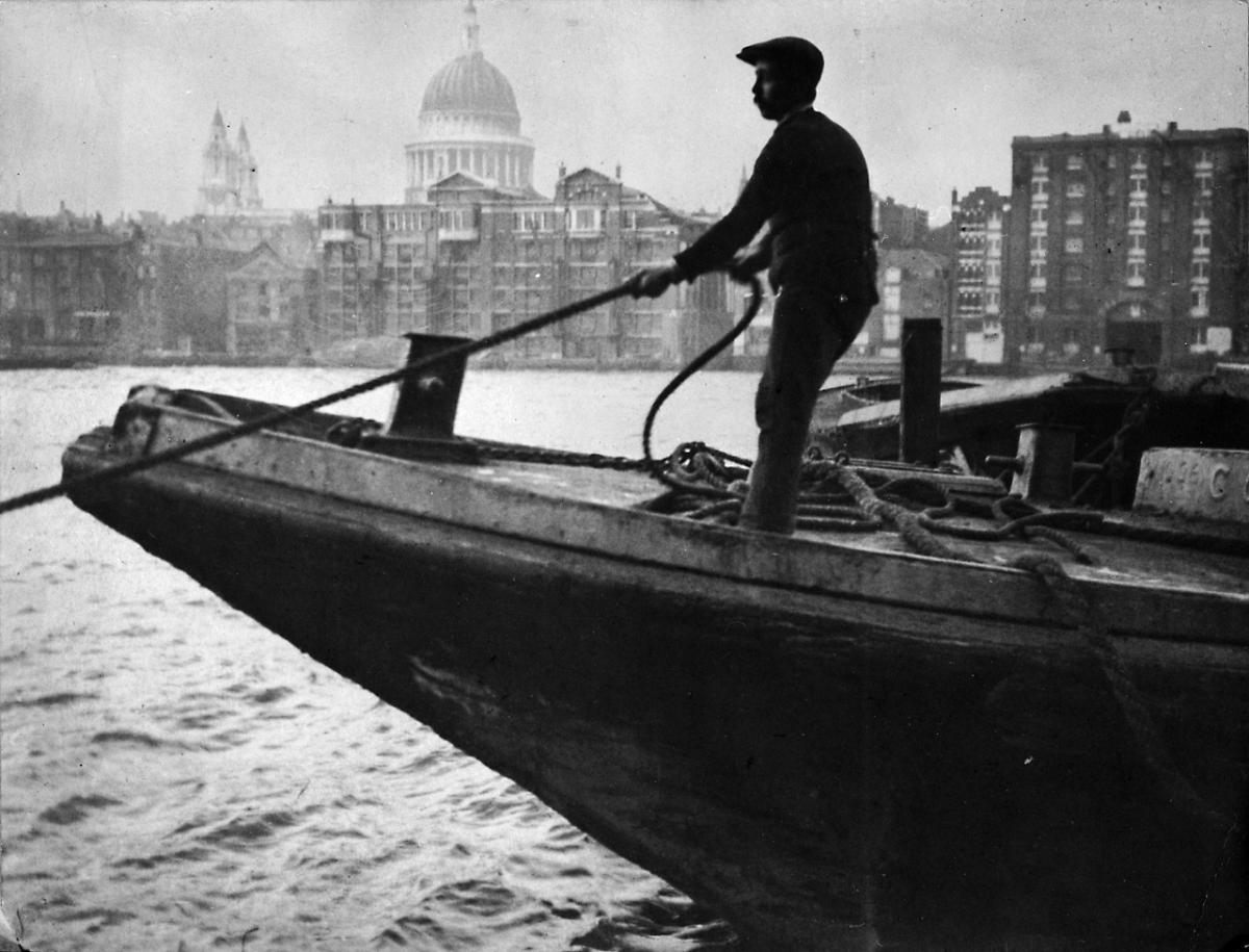 Historic photograph showing a man pulling on a rope on board a boat on the River Thames, with St Paul's Cathedral in the background