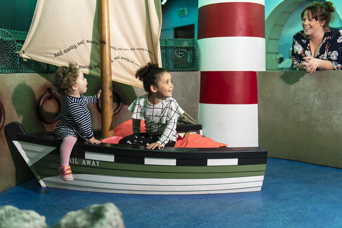 Two children play on a model boat in the National Maritime Museum's interactive AHOY Gallery, with a parent looking on