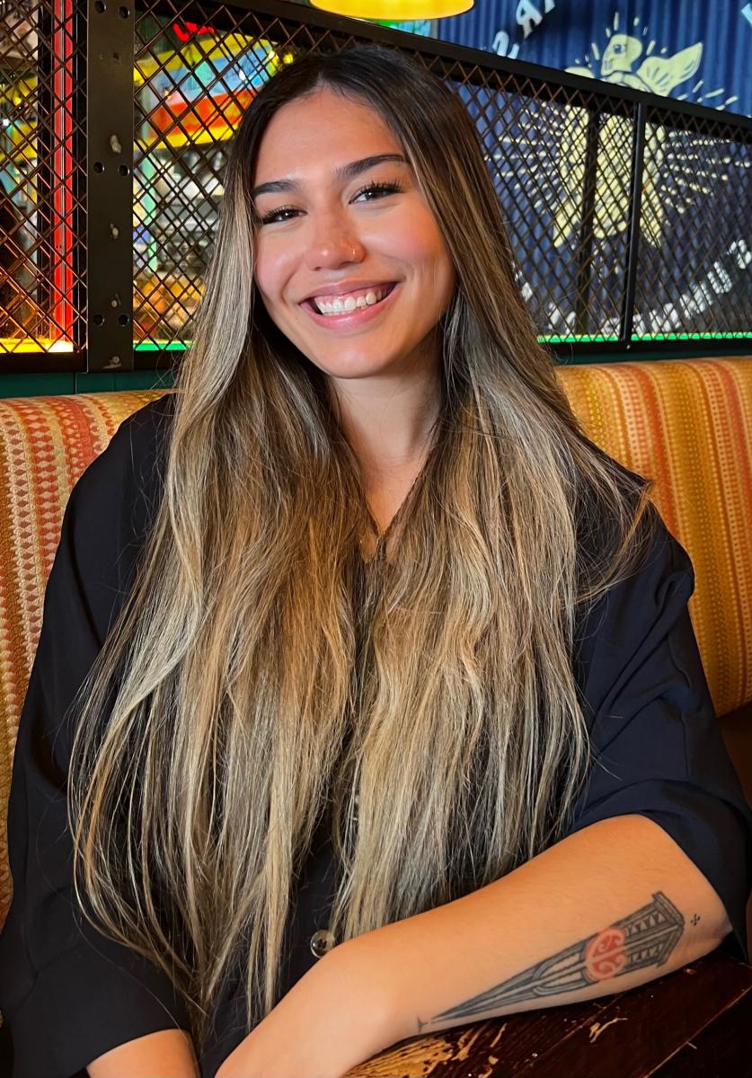 A portrait photograph of a young person with long sandy brown hair. They are smiling brightly at the camera, and a tattoo is visible on their left forearm
