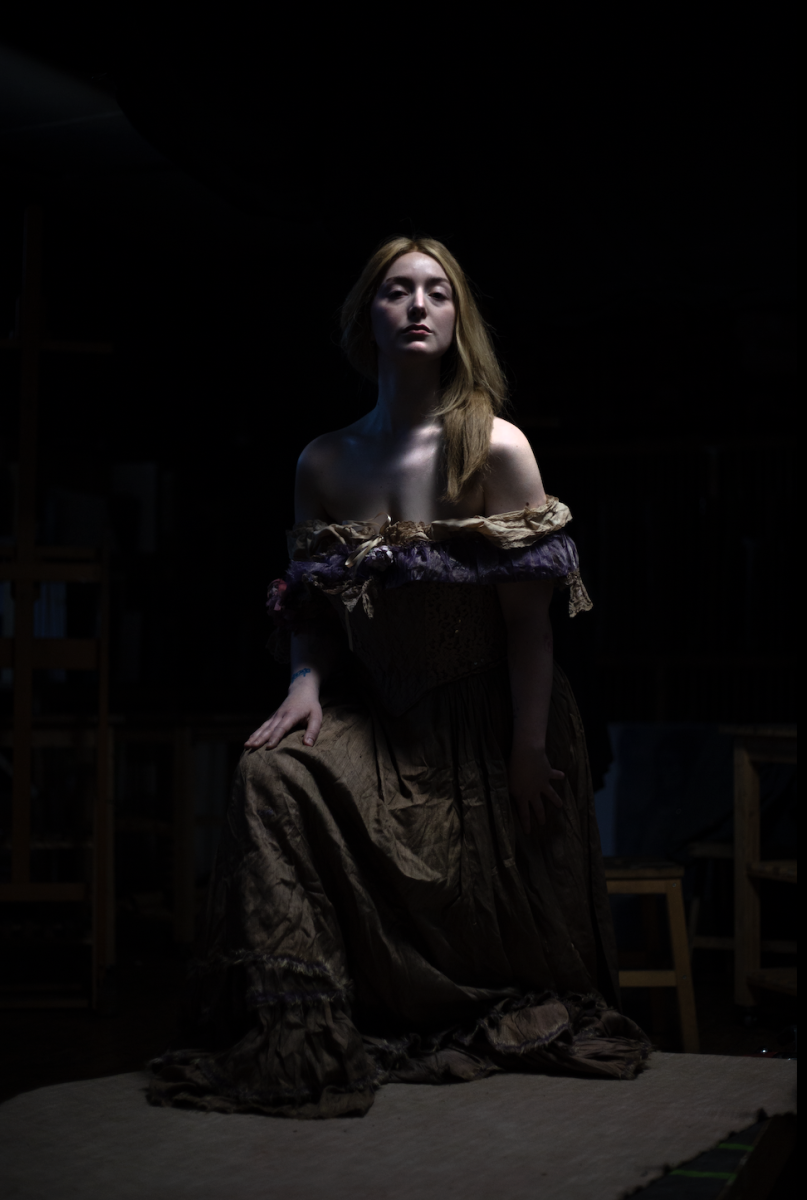 A photo of an actor playing Emma Hamilton. They kneel in the centre of a black background with long red hair falling over their shoulders. They wear a typical 19th century dress off the shoulders.