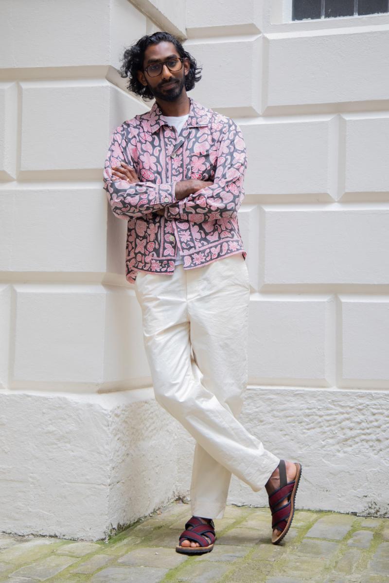 A portrait photograph of an Asian man in stylish pink shirt and white trousers. He is leaning nonchalantly against a white brick wall