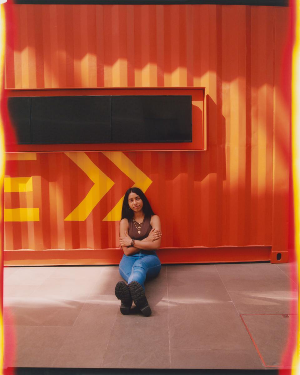 Photograph of a woman sitting on the ground next to an orange shipping container. She has her arms folded and is leaning against the corrugated metal