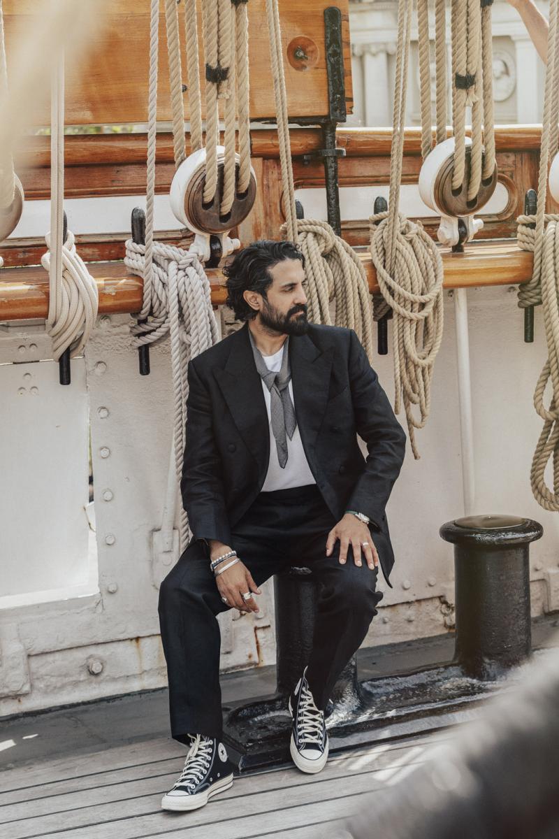 A man sits on the deck of a historic sailing ship, with ropes and rigging behind him. He wears a dark grey suit and black and white sneakers