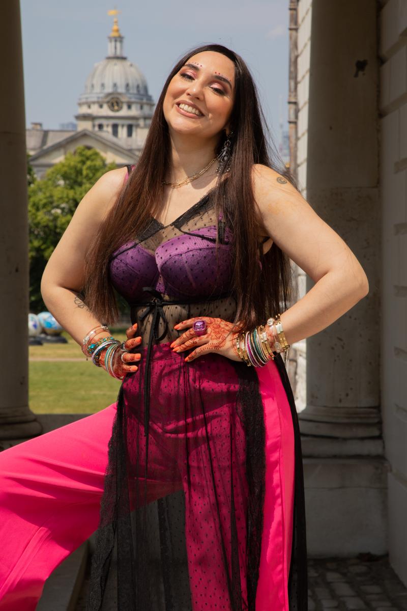 A photo of Namalee Bolle standing in the Queen's House colonnade with the Old Royal Naval College in the background. Namalee stands with her hands on her hips, smiling. She has long dark, stright hair that falls over her shoulders, is wearing a purple top, pink trouser and a black translucent tunic.