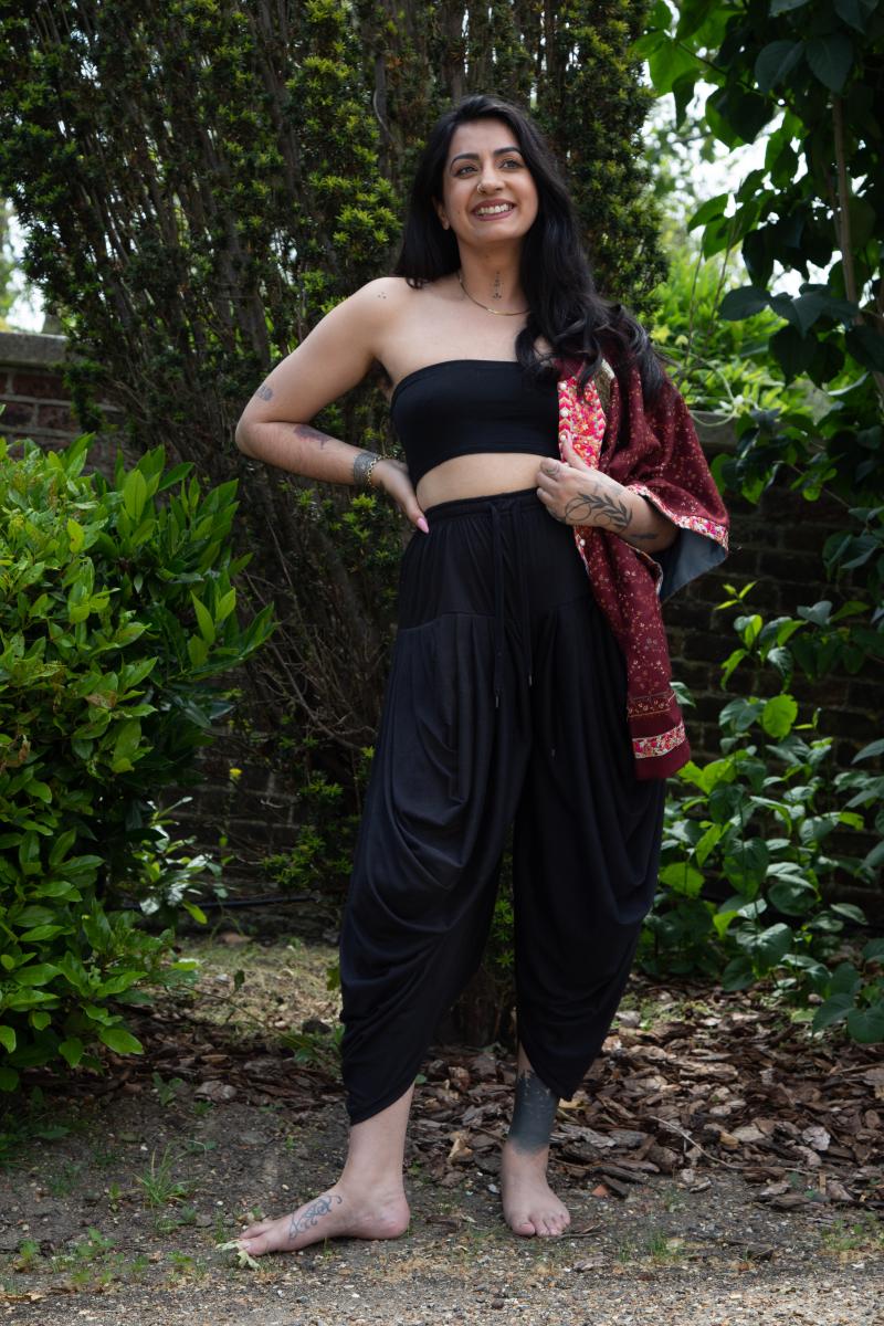 A photo of Sharan Dhaliwal who stands in front of vibrant greenery, wearing a black cropped top and high waisted black cropped trousers and a pink dupatta over one shoulder