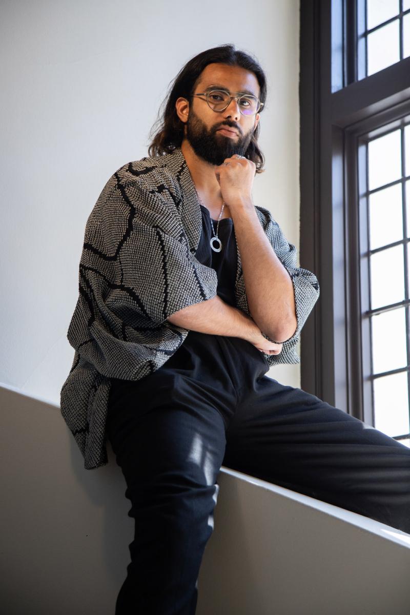 Portrait photograph of a man straddling a low wall. He has brown skin, a trimmed black beard and long dark hair