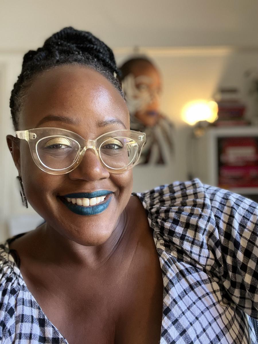 A photo of artist, Sharon Walters, who is Black woman smiling with blue lipstick and wearing translucent framed glasses and a gingham dress. Her hair is braided and tied up.