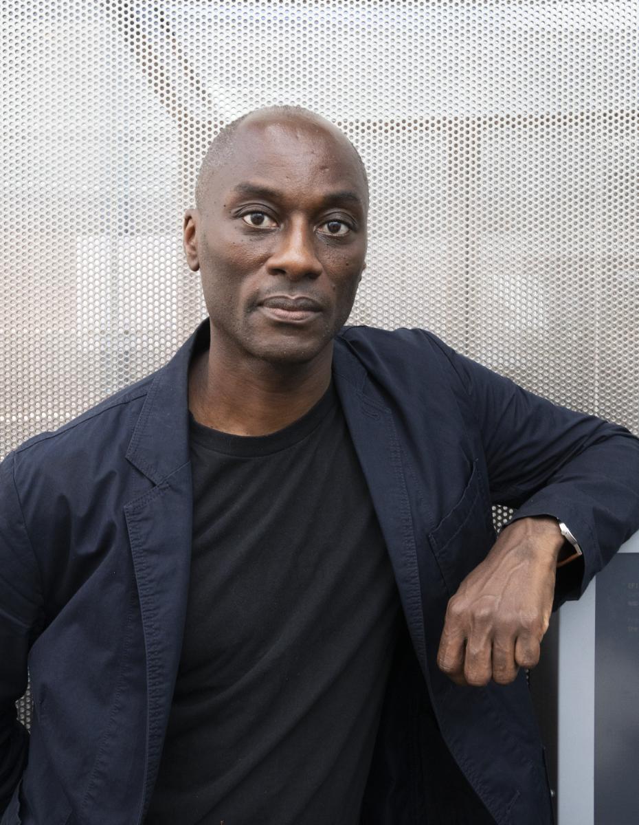 A photo of Ekow Eshun who stands casually with his arm resting on a tall plinth. He wears a dark blue blazer and black t-shirt.